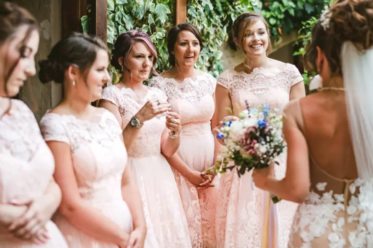 Bridesmaids in blush lace dresses smiling at Amie as she holds her bouquet