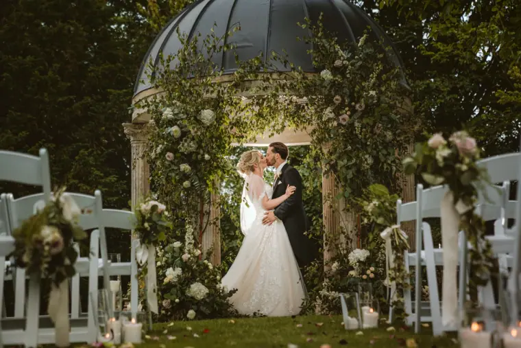 Abby and Taz kissing beneath a domed garden gazebo lined with white chairs at Ardington House