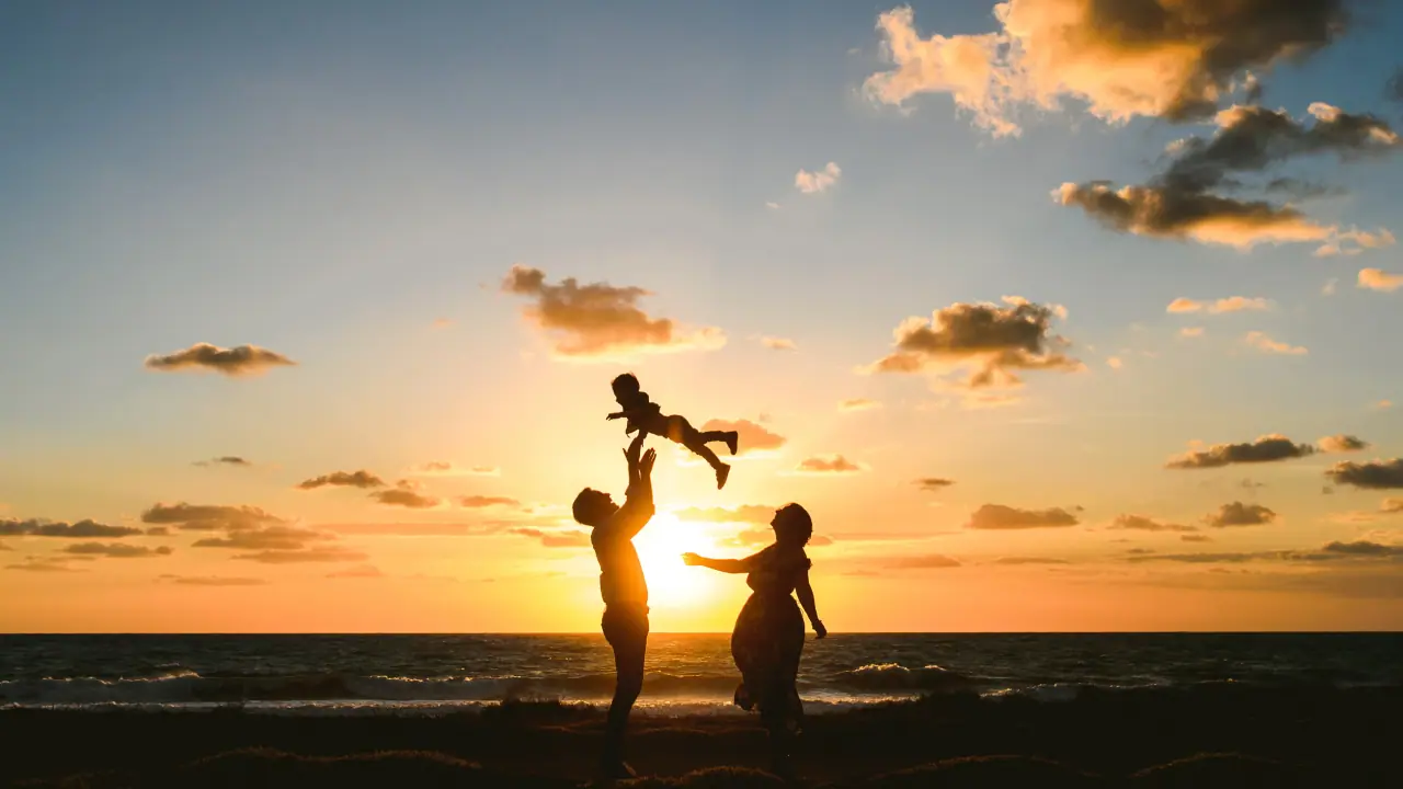 Ellie and James silhouetted against a seaside sunset, lifting their baby into the air