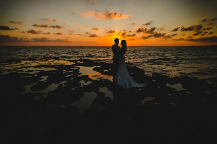 Bride and groom standing on dark coastal rocks under a golden sunset sky, Paphos