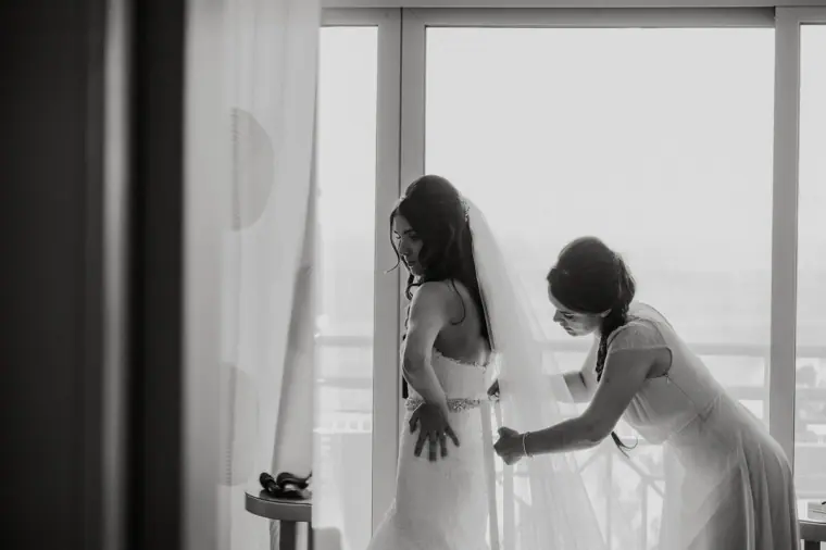 Black-and-white photo of a bridesmaid fastening Claire's dress by the window