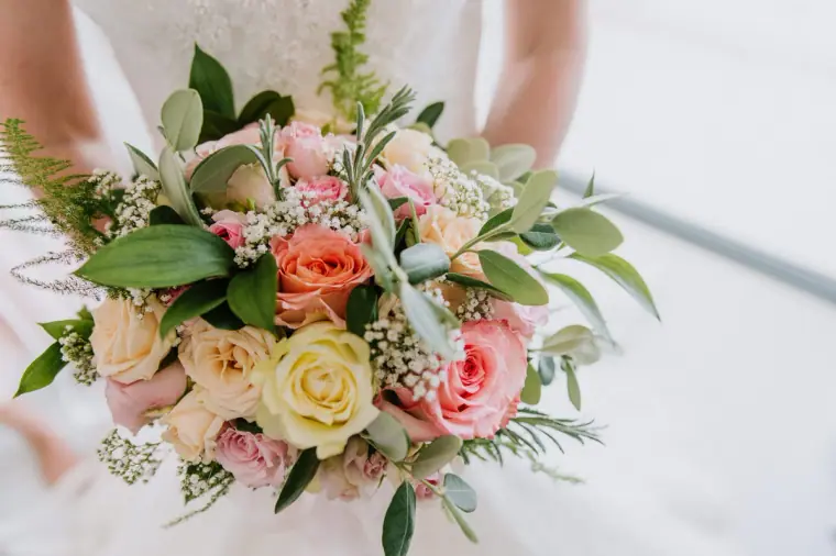 Peach, pink and yellow rose bouquet with olive leaves held against a white dress