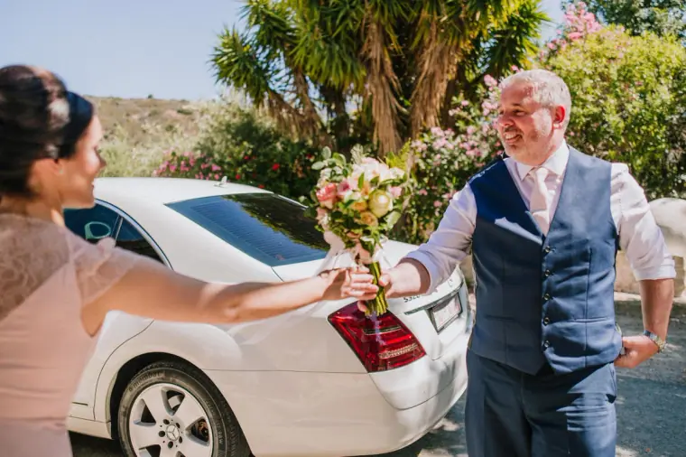 Bridesmaid handing a bouquet to Claire's father beside the white wedding car