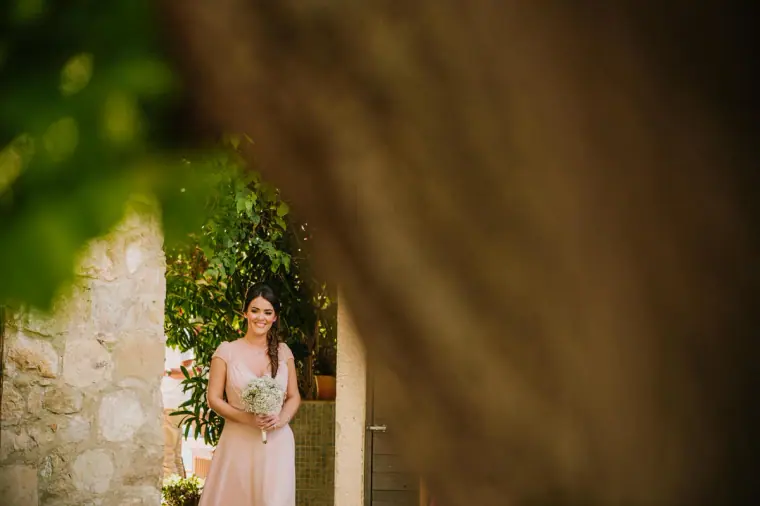 Bridesmaid with a bouquet waiting by a stone wall, framed by a tree