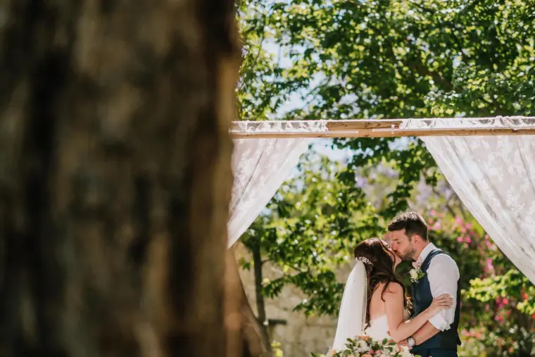 Claire and Sean kissing beneath the lace arch, framed by a tree trunk