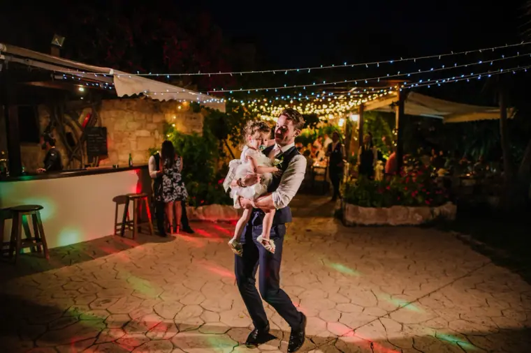 Man in a waistcoat dancing with a toddler on the lit dance floor at night