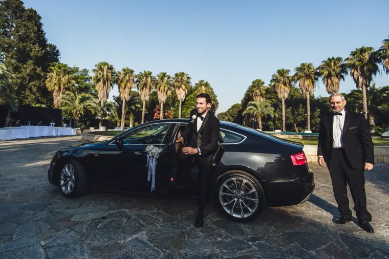 Angelos stepping out of a black car on a palm-lined avenue