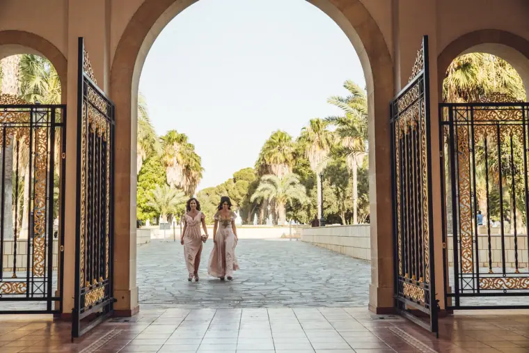 Two bridesmaids walking through an ornate gateway towards a palm-lined avenue