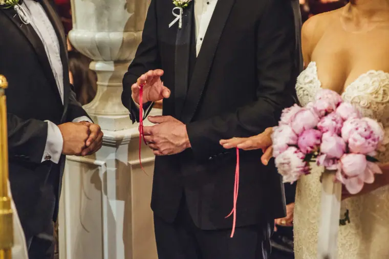 Close-up of the groom's hands with a red ribbon beside the bride's peonies