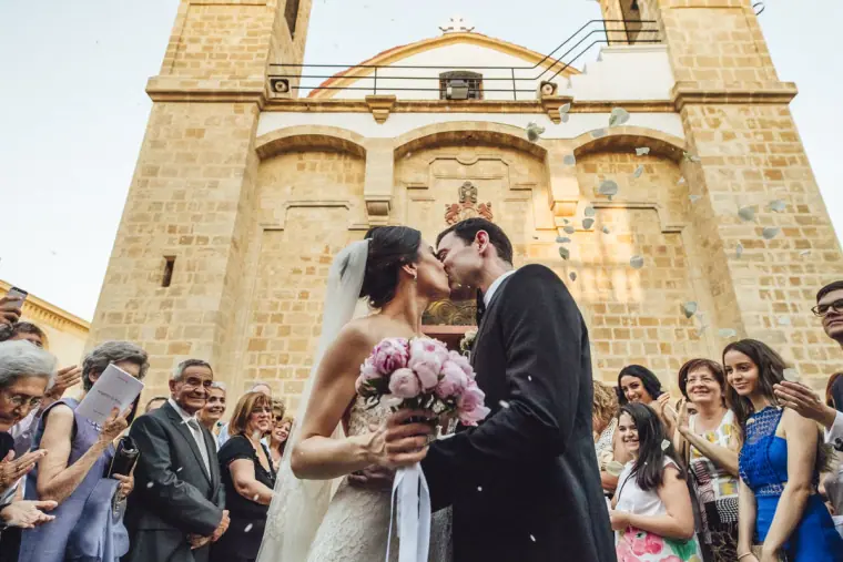 Ruby and Angelos kissing outside the stone church as guests cheer, peony bouquet in hand