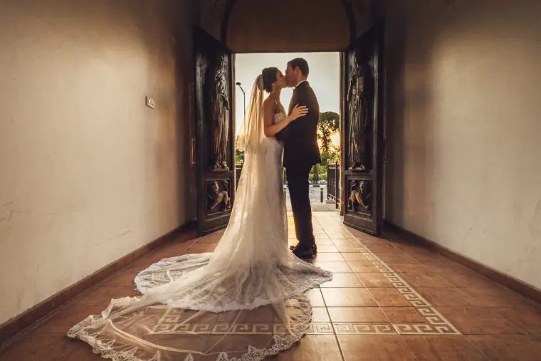 Ruby and Angelos kissing in a sunlit doorway, her lace train fanned on the tiled floor
