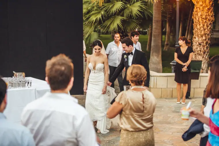 Bride and groom arriving through the palm-lined reception garden past guests