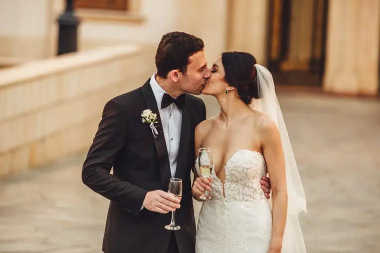 Ruby and Angelos sharing a kiss, champagne glasses in hand, on a stone terrace