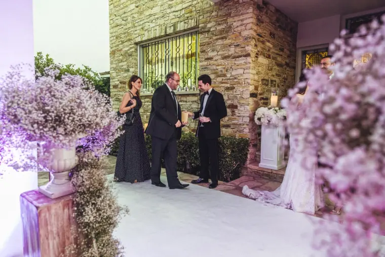 Ruby and Angelos greeting guests beside lilac gypsophila arrangements at a stone house