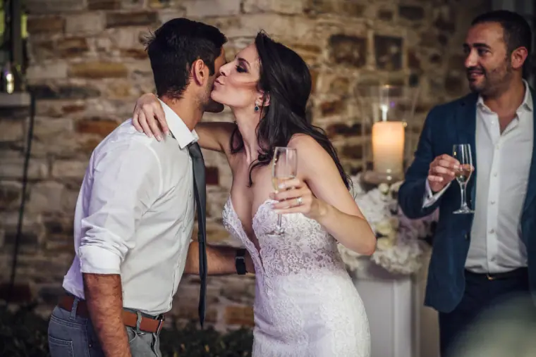 Bride kissing a guest on the cheek while the groom raises a glass by a stone wall