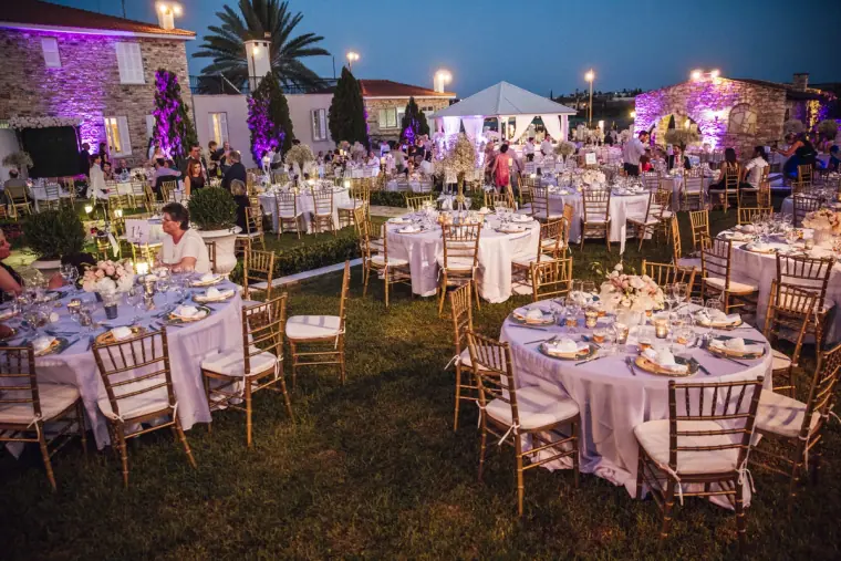 Reception lawn of round tables with gold chairs beneath purple-lit trees at dusk