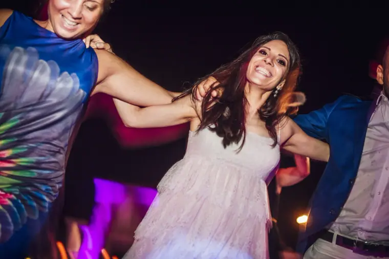 Bride dancing joyfully with friends in a floral dress under purple light