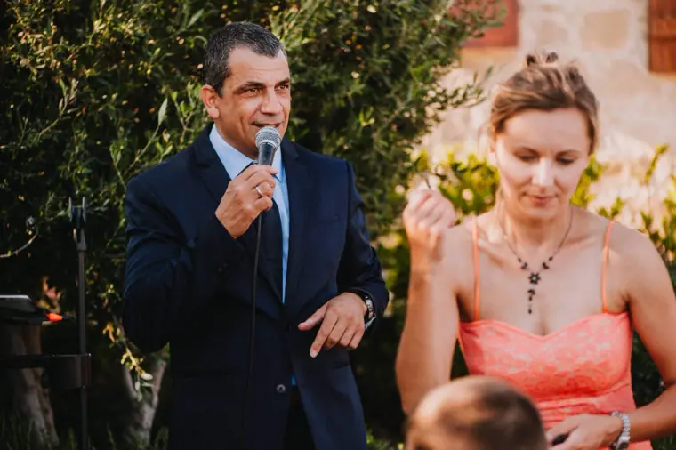 Guest giving a speech into a microphone beside a bridesmaid in coral at Liopetro, Kouklia