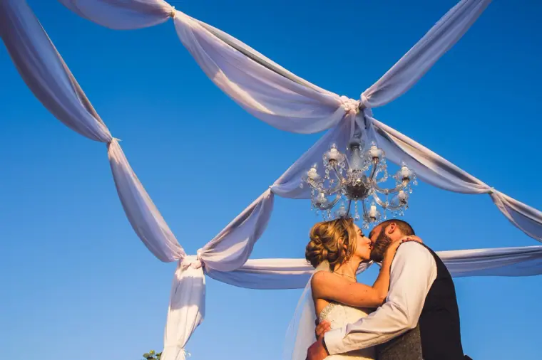 Bride and groom kissing beneath a chandelier and white draped fabric against a blue sky at Liopetro