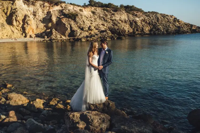 Bride and groom standing on rocks at the edge of a calm sea cove beneath cliffs, Elixir, Ibiza