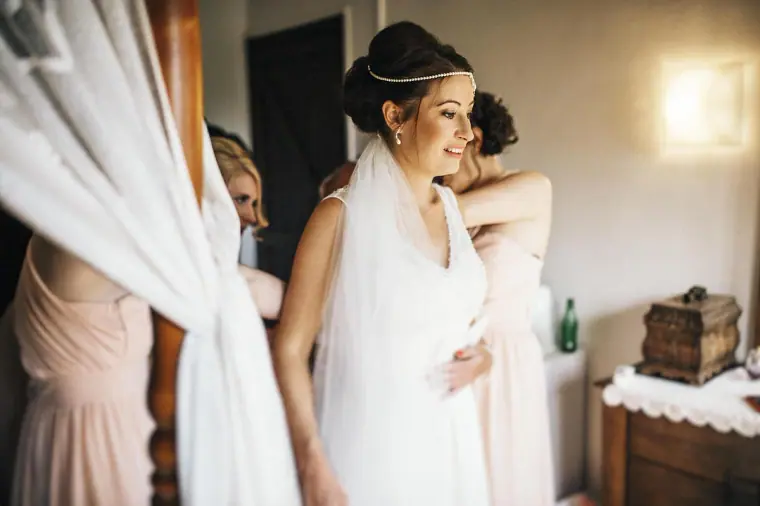 Bride smiling as her dress is fastened beside white curtains