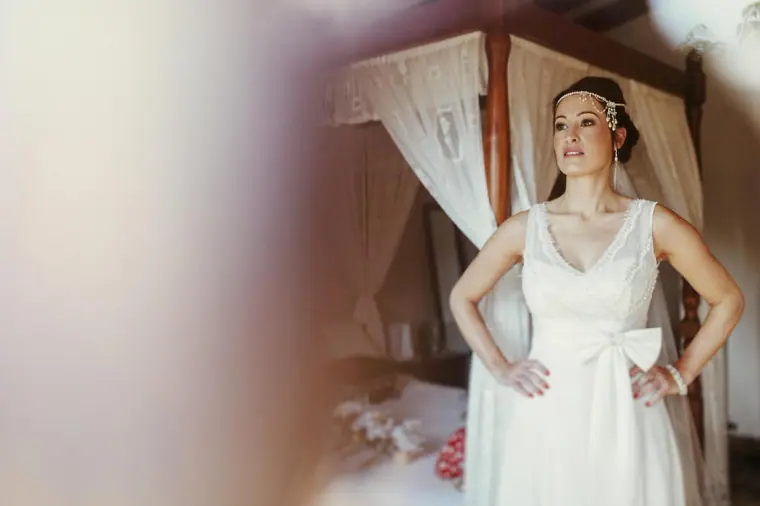 Donna standing by a canopy bed in her bow-backed wedding dress and pearl headpiece