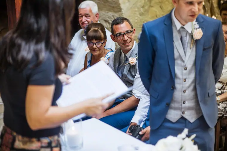 Groom in a blue suit and grey waistcoat smiling as the registrar reads the ceremony