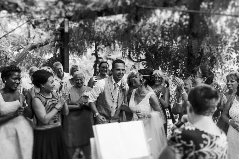 Black-and-white photo of guests clapping and laughing under the vine-covered pergola