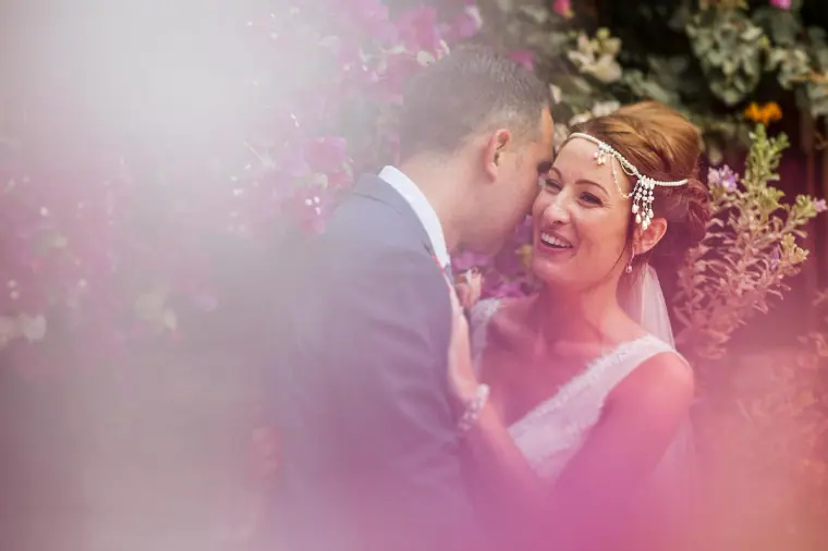 Bride smiling as the groom kisses her cheek, framed by soft pink blossom