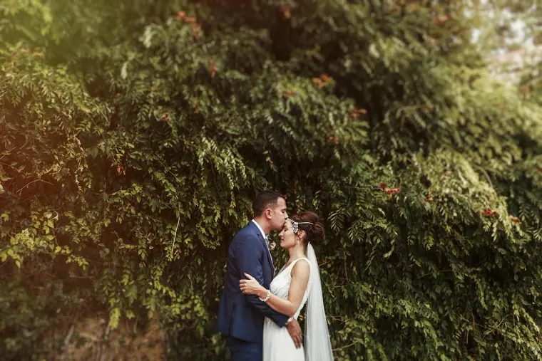Byron kissing Donna's forehead in front of a dense wall of greenery
