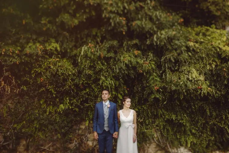 Bride and groom standing side by side before tall green foliage