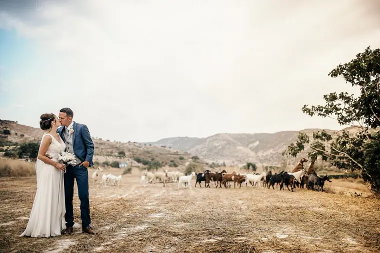 Couple standing in a dusty field as a herd of goats grazes behind them