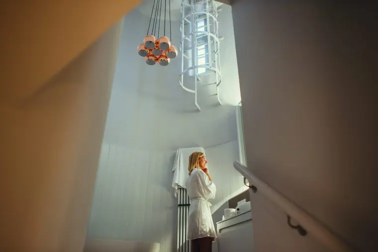 Bridesmaid in a white robe standing on a spiral staircase beneath a pendant light