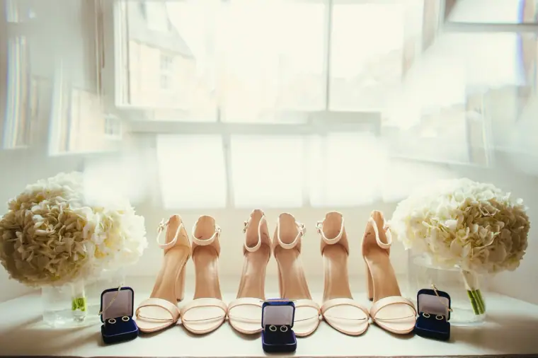 Row of blush sandals and ring boxes on a sunlit window ledge between white rose bouquets