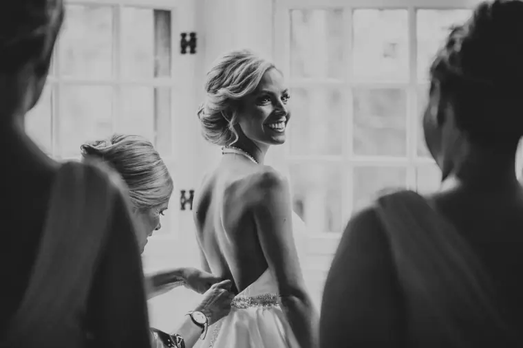 Black-and-white photo of the bride smiling as bridesmaids help her into her dress