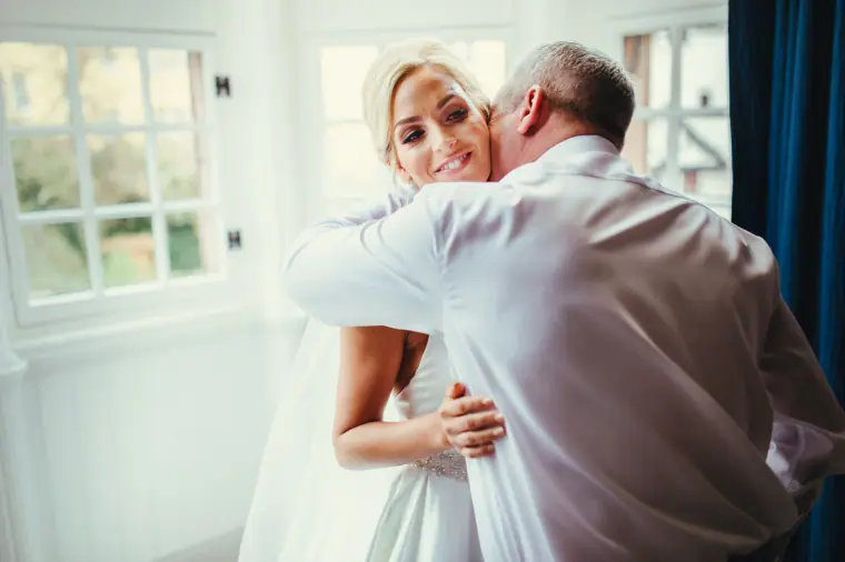 Bride smiling over her father's shoulder as he hugs her by the window