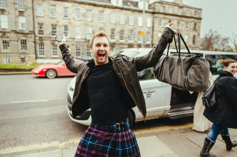 Guest in a tartan kilt cheering with a holdall on an Edinburgh street