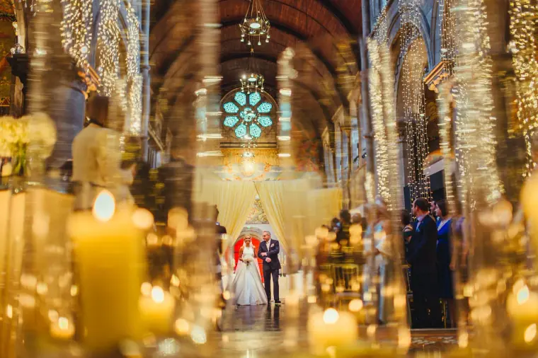 Bride and father walking up the aisle beneath a rose window, framed by candles