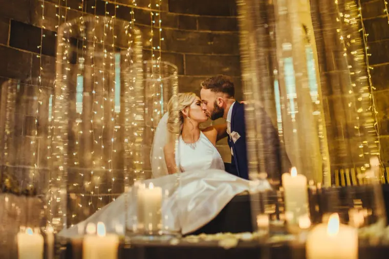 Bride and groom kissing as she sits among candles and curtains of fairy lights