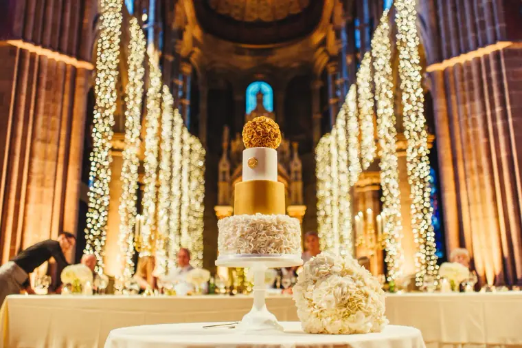 Gold-and-white wedding cake on a table before the lit nave of Mansfield Traquair