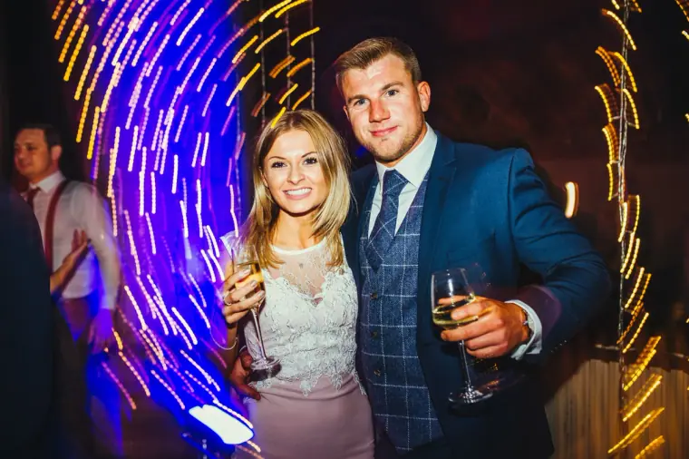 Smiling couple of guests posing with drinks under swirling purple lights