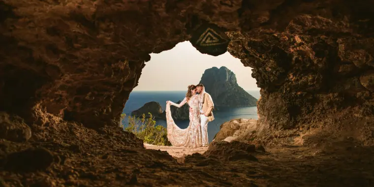 Bride and groom framed by a rocky cave mouth overlooking the sea and a rocky islet