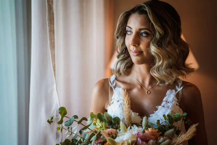 Bride gazing out of a window beside her pastel bouquet in soft light