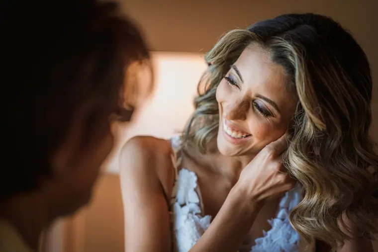 Smiling bride touching her hair while getting ready, seen past a friend