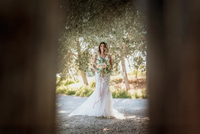 Bride with her bouquet among olive trees, framed through a dark opening