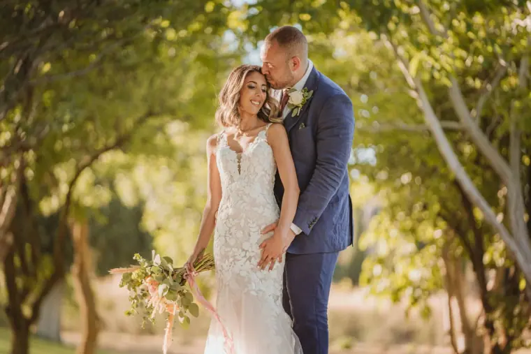 Groom kissing Emily's temple among sunlit trees at Minthis, Paphos