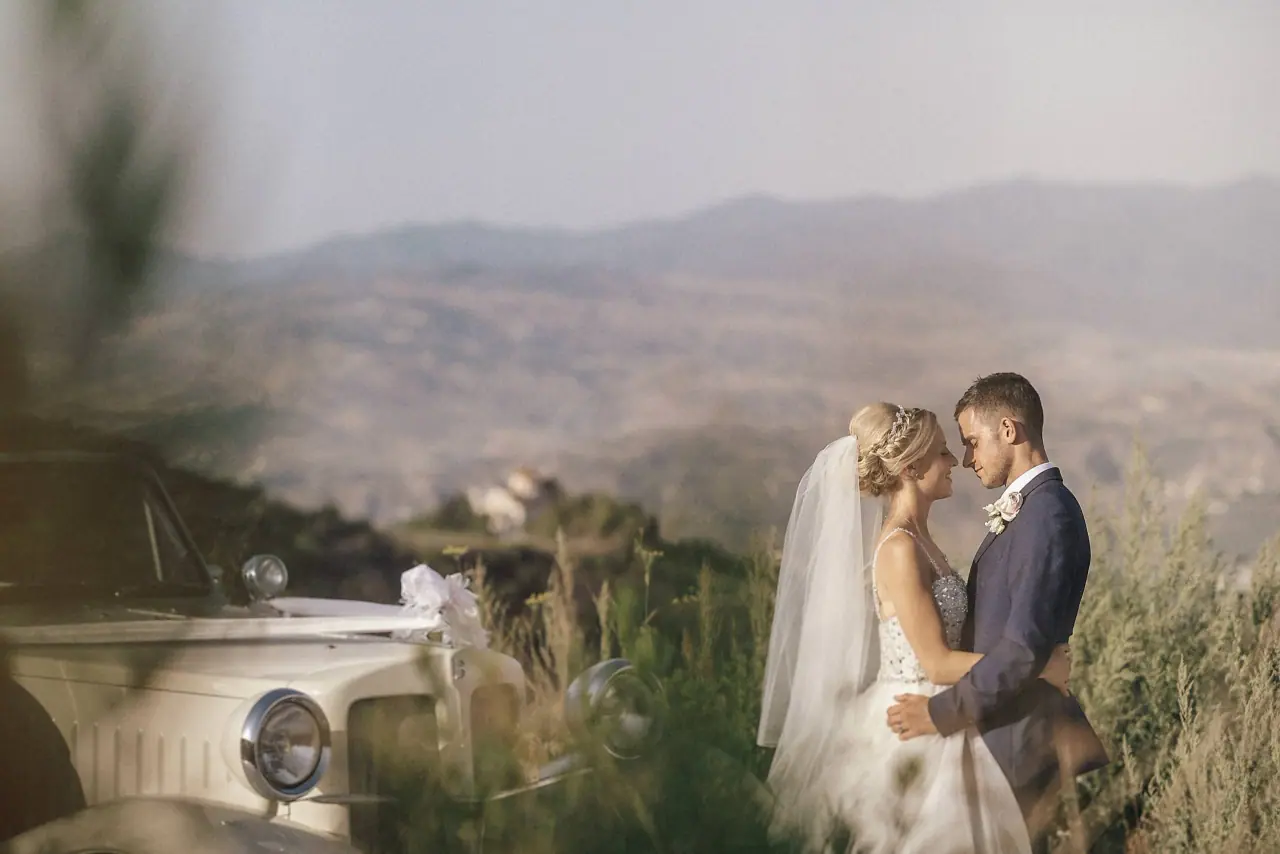 Emmee and Danny embracing beside a vintage car above the hills at Ayii Anargyri