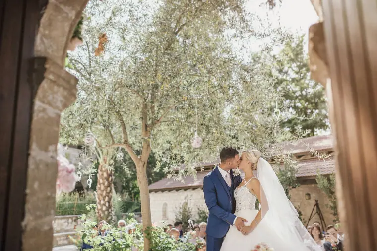 Bride and groom kissing under olive trees, framed by a stone arch