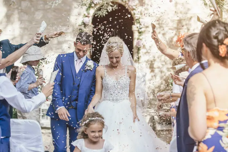 Bride and groom laughing under a shower of confetti at the stone chapel