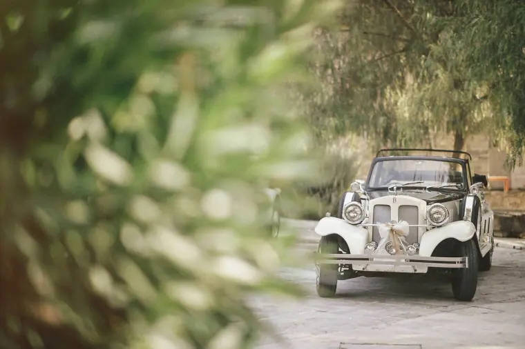 Vintage white wedding car parked on the drive, framed by blurred foliage